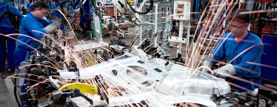 Zwei Männer in blauer Arbeitskleidung arbeiten in einer Produktionshalle mit einem Schweißgerät, Funken fliegen.