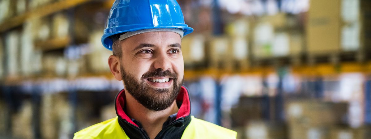 Lagermitarbeiter steht mit Schutzkleidung und Helm vor einem Hochregal und lächelt.