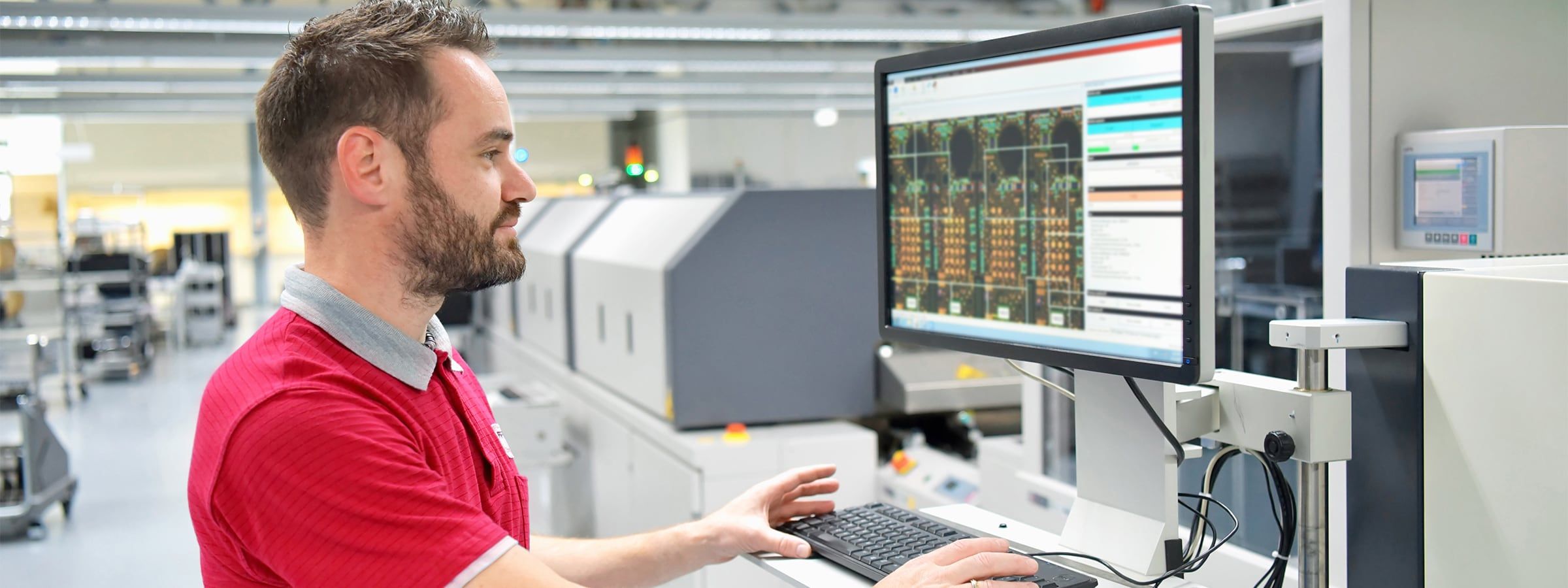 Maschinenbediener in rotem Poloshirt arbeitet an einem Industriecomputer in einer modernen Produktionshalle. Auf dem Bildschirm sind technische Zeichnungen oder Schaltpläne zu sehen. Im Hintergrund befinden sich automatisierte Maschinen und Förderanlagen. Das Bild vermittelt technisches Know-how, Präzision und Digitalisierung in der industriellen Fertigung.