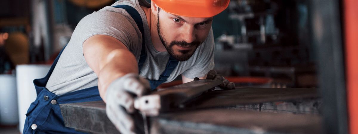 Metallbauer in blauer Arbeitskleidung und orangefarbenem Schutzhelm konzentriert sich auf das Bearbeiten eines Metallstücks in einer Werkstatt.