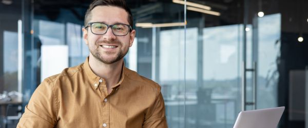 Ein lächelnder Mann mit Brille und braunem Hemd sitzt an einem Schreibtisch mit einem Laptop in einem modernen Büro.