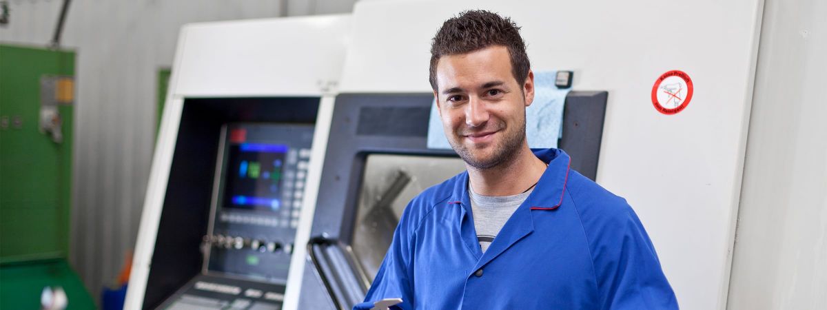 Freundlicher Maschinenbediener in blauer Arbeitsjacke steht vor einer CNC-Maschine in einer Werkhalle. Er hält ein Bauteil in der Hand und blickt lächelnd in die Kamera. Im Hintergrund sind Bedienelemente und industrielle Ausstattung sichtbar. Das Bild vermittelt Fachwissen, Präzision und eine positive Arbeitsatmosphäre im Bereich Maschinenbedienung oder Metallverarbeitung.