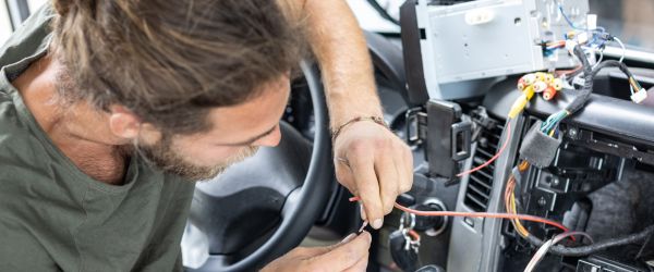 Ein Kfz-Mechatroniker arbeitet konzentriert an der Verkabelung im Inneren eines Autos, um technische Komponenten zu verbinden.