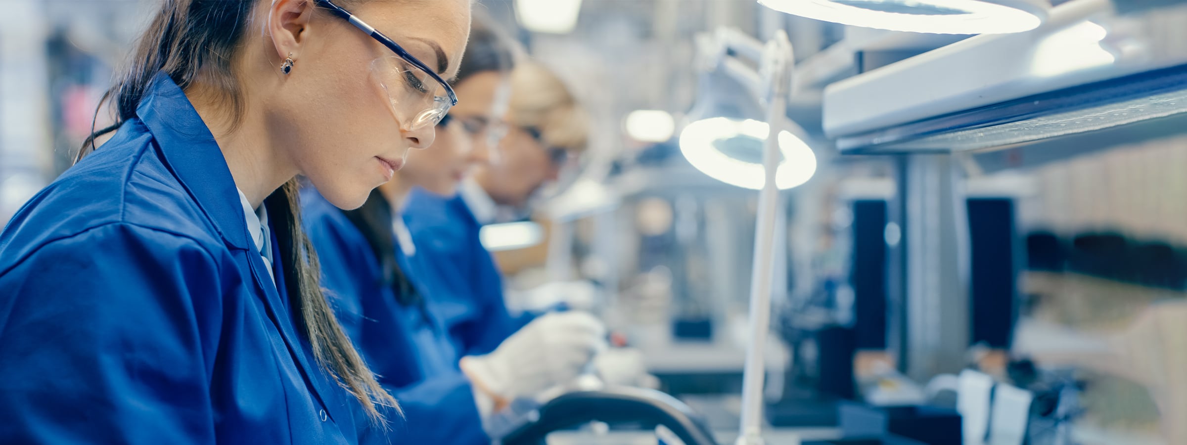 Frauen in blauen Arbeitskitteln und Schutzbrillen arbeiten konzentriert an elektronischen Bauteilen in einem Labor.