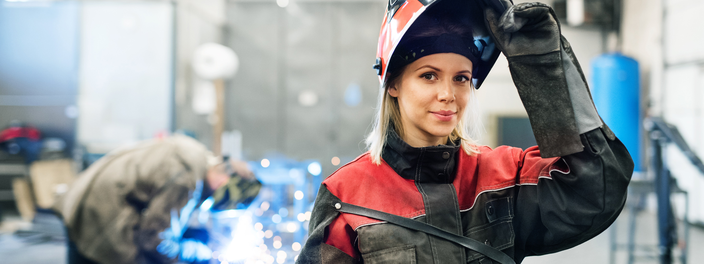 Eine Frau in Schutzkleidung hebt ihren Schweißhelm leicht an, während im Hintergrund eine Person an einer Metallwerkbank arbeitet.