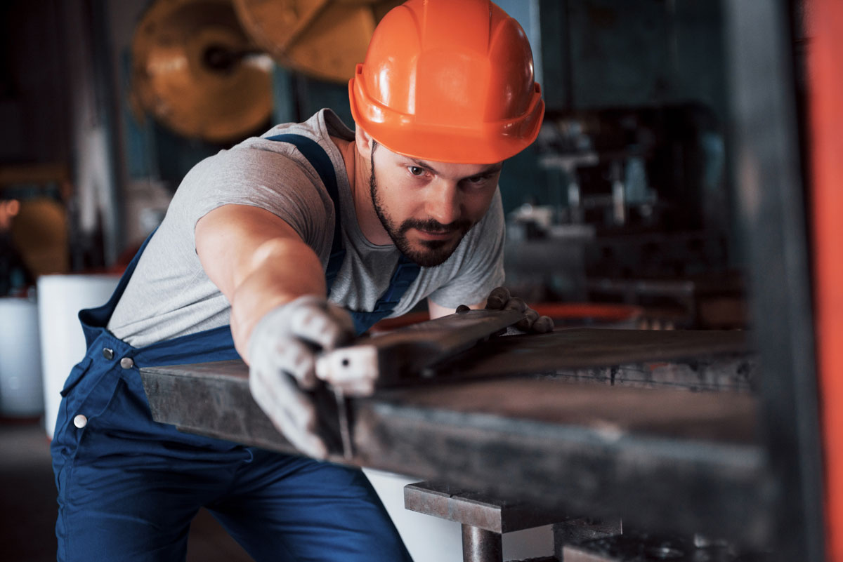 Metallbauer in blauer Arbeitskleidung und orangefarbenem Schutzhelm konzentriert sich auf das Bearbeiten eines Metallstücks in einer Werkstatt.