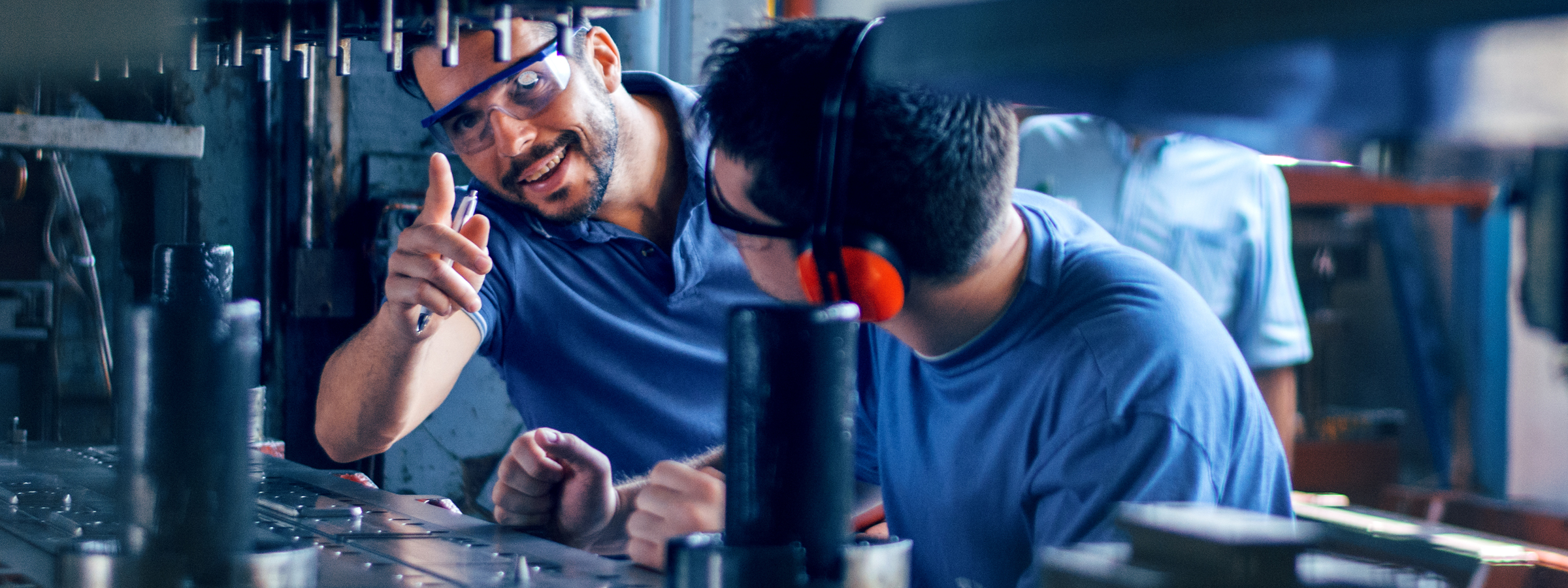 Zwei Arbeiter in einer Metallwerkstatt, die Sicherheitsbrillen tragen. Der eine erklärt dem anderen lächelnd etwas an einer Maschine.