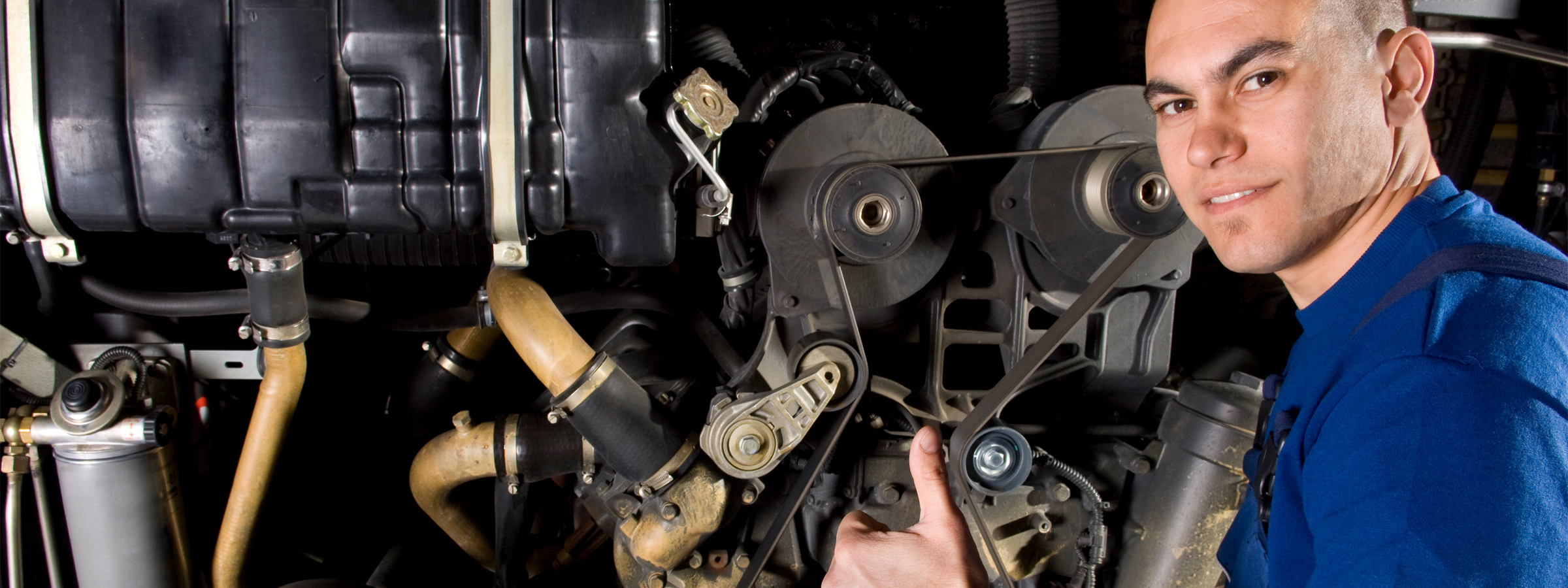 Ein Mechaniker in blauer Arbeitskleidung arbeitet an einem Motor und zeigt mit einer Daumen-hoch-Geste in die Kamera.
