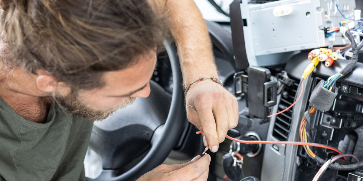 Ein Kfz-Mechatroniker arbeitet konzentriert an der Verkabelung im Inneren eines Autos, um technische Komponenten zu verbinden.