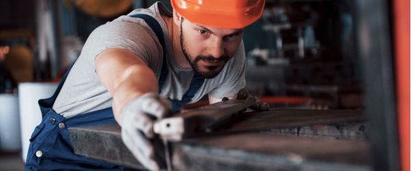 Metallbauer in blauer Arbeitskleidung und orangefarbenem Schutzhelm konzentriert sich auf das Bearbeiten eines Metallstücks in einer Werkstatt.