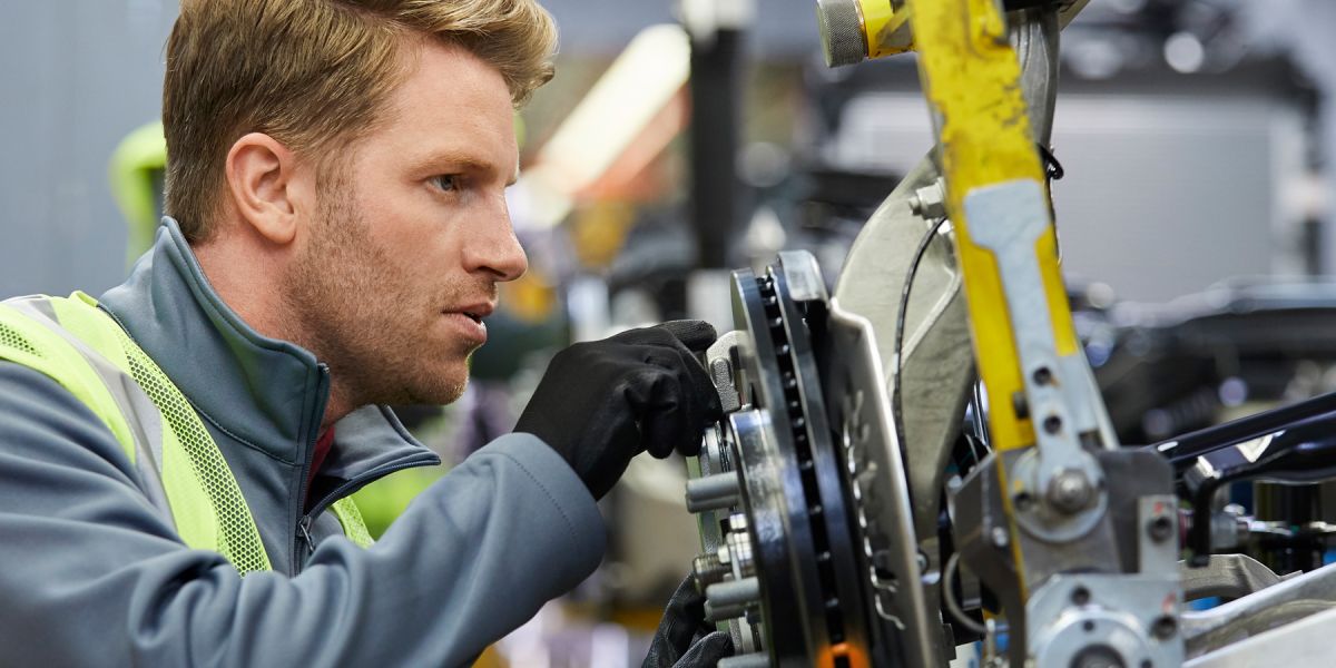 Ein Industriemechaniker in Warnweste und Handschuhen arbeitet konzentriert an einer Maschine in einer Produktionshalle.