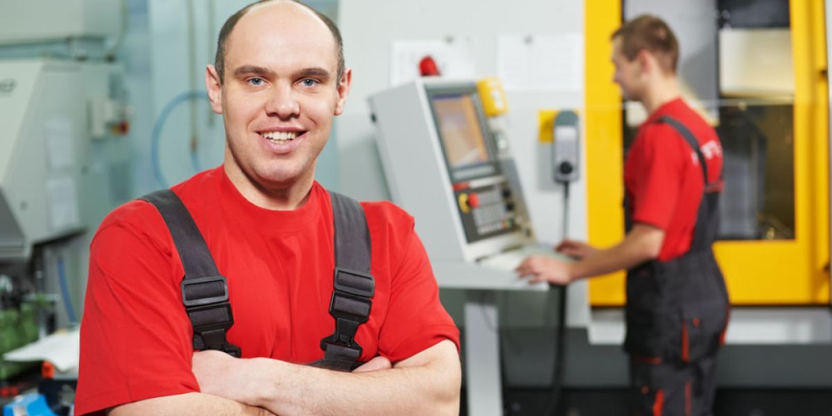 Ein lächelnder Arbeiter in rotem Shirt steht mit verschränkten Armen in einer Werkstatt, während im Hintergrund jemand eine Maschine bedient.