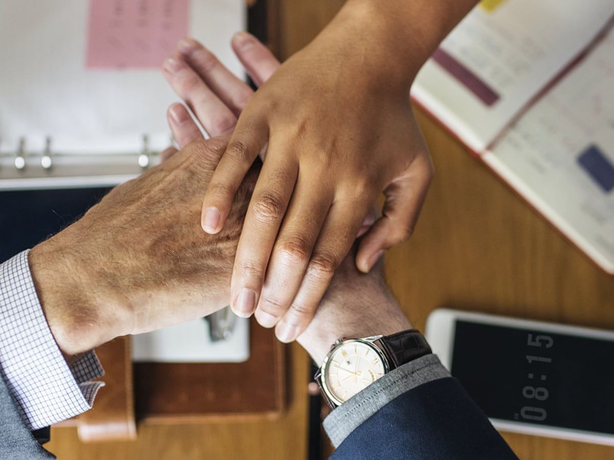 Mehrere Hände werden aufeinandergelegt, als Zeichen der Zusammenarbeit, auf einem Schreibtisch mit Unterlagen und Smartphone.