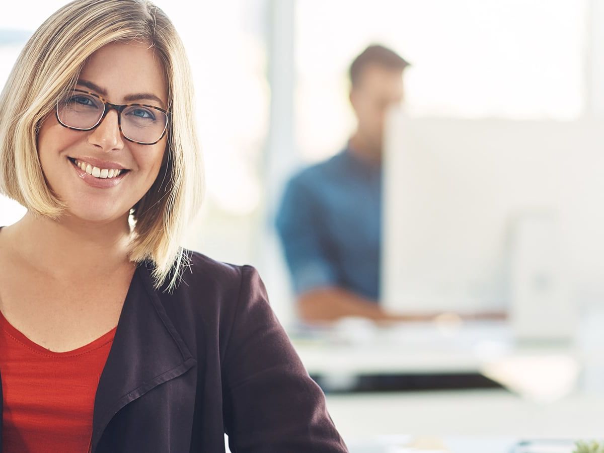 Eine lächelnde Frau mit Brille sitzt in einem Büro, während im Hintergrund eine Person an einem Computer arbeitet.