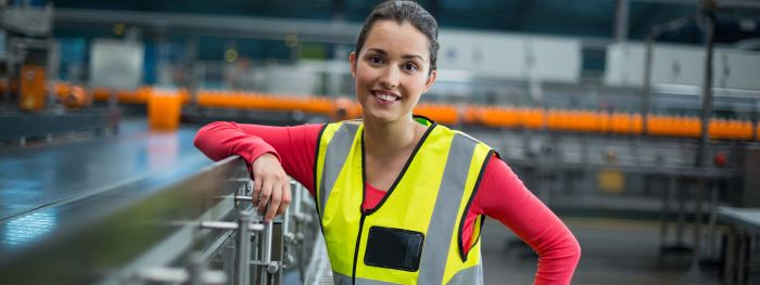 Eine Fliessbandarbeiterin in Warnweste lehnt sich lächelnd an ein Geländer in einer Produktionshalle.