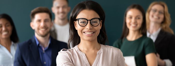 Eine Frau mit Brille lächelt im Vordergrund, während eine Gruppe von Menschen im Hintergrund ebenfalls lächelt.