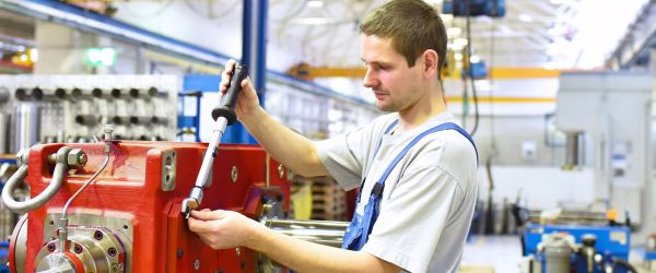 Monteur bei der Maschinenmontage in einer Industriehalle: Ein Mann in Arbeitskleidung mit blauer Latzhose zieht konzentriert eine Schraube an einem großen, roten Maschinenteil mit einem Drehmomentschlüssel fest. Im Hintergrund sind weitere Maschinen, Werkzeuge und Bauteile sichtbar. Die Szene vermittelt Präzision, technisches Know-how und handwerkliche Tätigkeit im industriellen Umfeld.