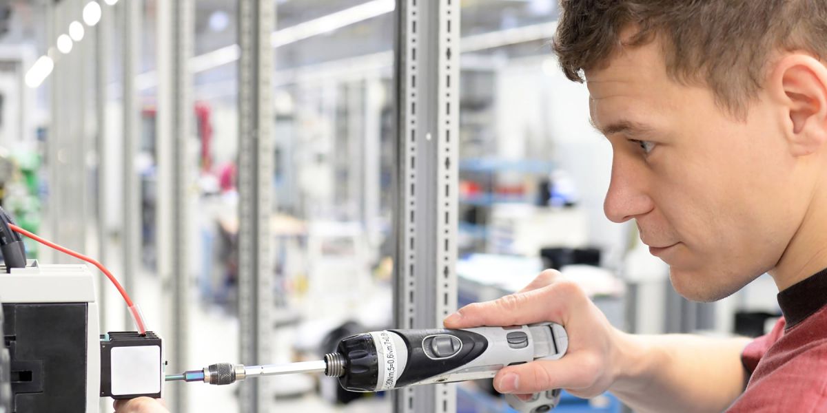 Ein lächelnder Mann mit Brille und braunem Hemd sitzt an einem Schreibtisch mit einem Laptop in einem modernen Büro.