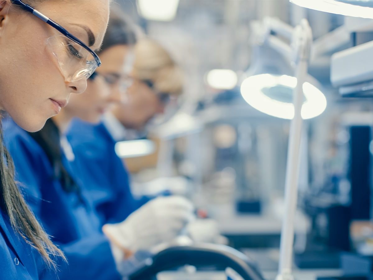 Frauen in blauen Arbeitskitteln und Schutzbrillen arbeiten konzentriert an elektronischen Bauteilen in einem Labor.