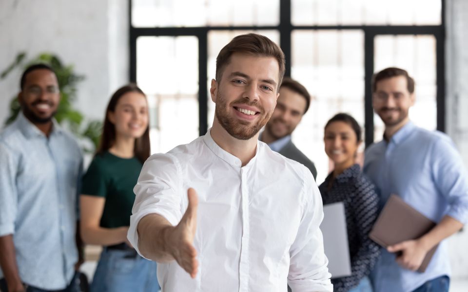 Ein lächelnder Mann streckt die Hand zum Händeschütteln aus, während im Hintergrund ein Team freundlich zuschaut.