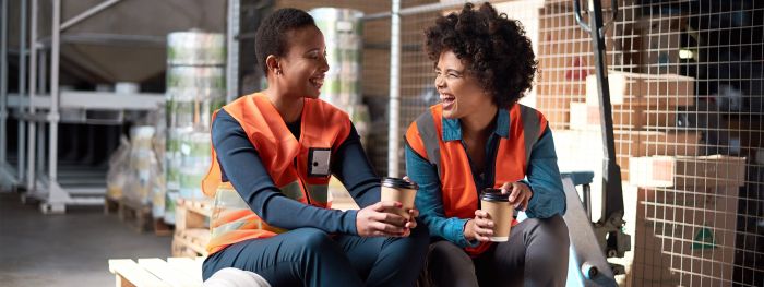 Zwei Lagerarbeiterinnen in Warnwesten sitzen lächelnd auf Paletten in einem Lager und halten Kaffeebecher in der Hand.