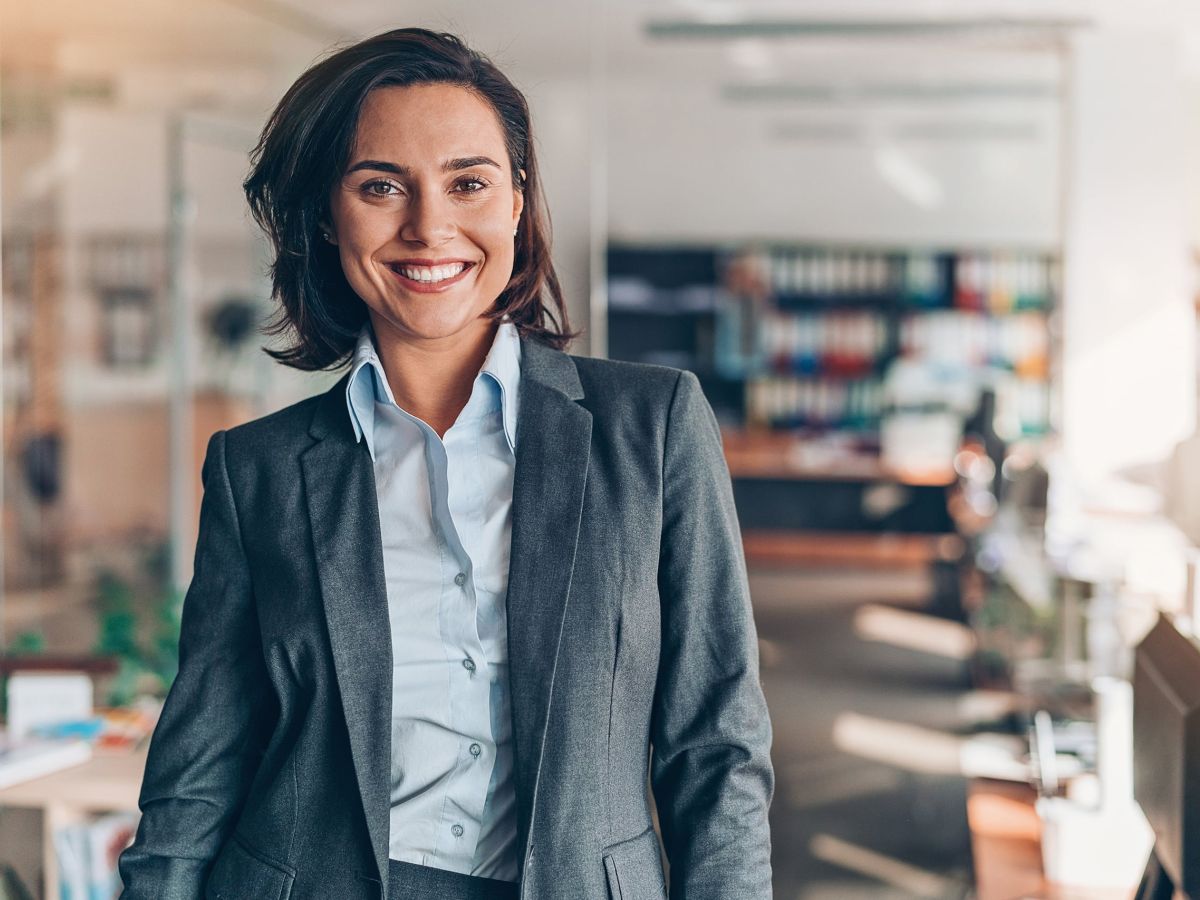 Eine lächelnde Geschäftsfrau in einem grauen Anzug steht in einem modernen Büro mit Glaswänden und Regalen im Hintergrund.