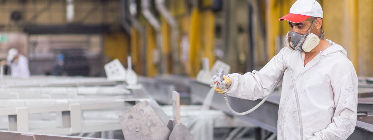 Lackierer im Schutzanzug mit Handschuhen und Maske lackiert mit einer Spritzpistole Metallteile in einer Industriehalle