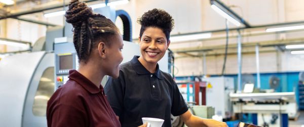 Zwei Frauen unterhalten sich lächelnd in einer modernen Industriehalle. Beide tragen Arbeitskleidung in Polohemden, eine hält einen Kaffeebecher. Im Hintergrund sind Maschinen und Werkbänke sichtbar. Das Bild vermittelt Teamwork, Vielfalt und ein positives Arbeitsklima in einem technischen oder industriellen Umfeld.