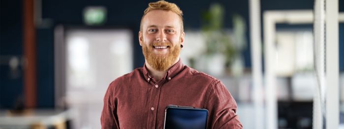 Ein lächelnder Mann mit Bart in einem roten Hemd hält ein Tablet in der Hand und steht in einem modernen Büro.