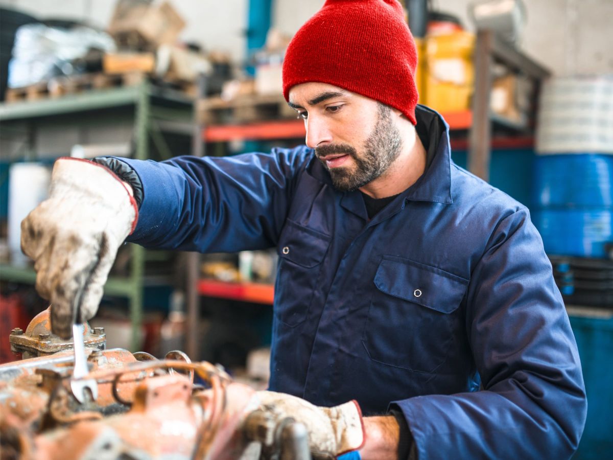 Ein Mechaniker in blauer Arbeitskleidung und roter Mütze arbeitet konzentriert mit einem Schraubenschlüssel an einer Maschine.