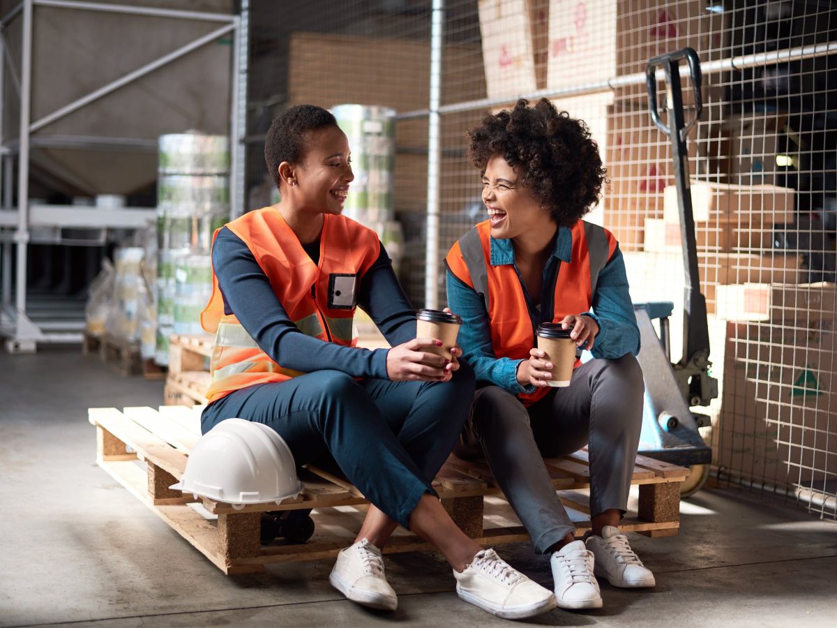 Zwei Lagerarbeiterinnen in Warnwesten sitzen lachend auf einer Palette in einem Lager und halten Kaffeebecher in der Hand.