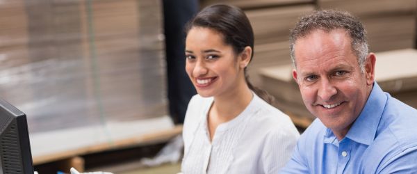 Zwei lächelnde Büromitarbeiter sitzen vor einem Computer und arbeiten in einer Lagerumgebung zusammen.