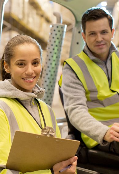 Zwei Lagerarbeiter in Warnwesten, die Frau hält ein Klemmbrett, beide lächeln in einem Lagerhaus.