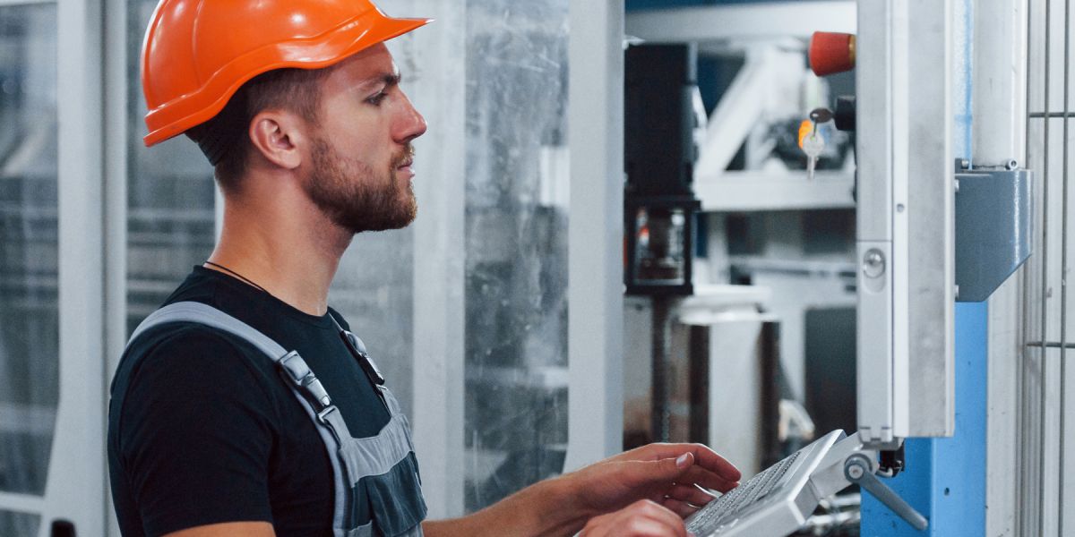 Ein Industriearbeiter mit orangem Schutzhelm bedient eine Maschine über ein Bedienfeld in einer automatisierten Produktionsanlage.