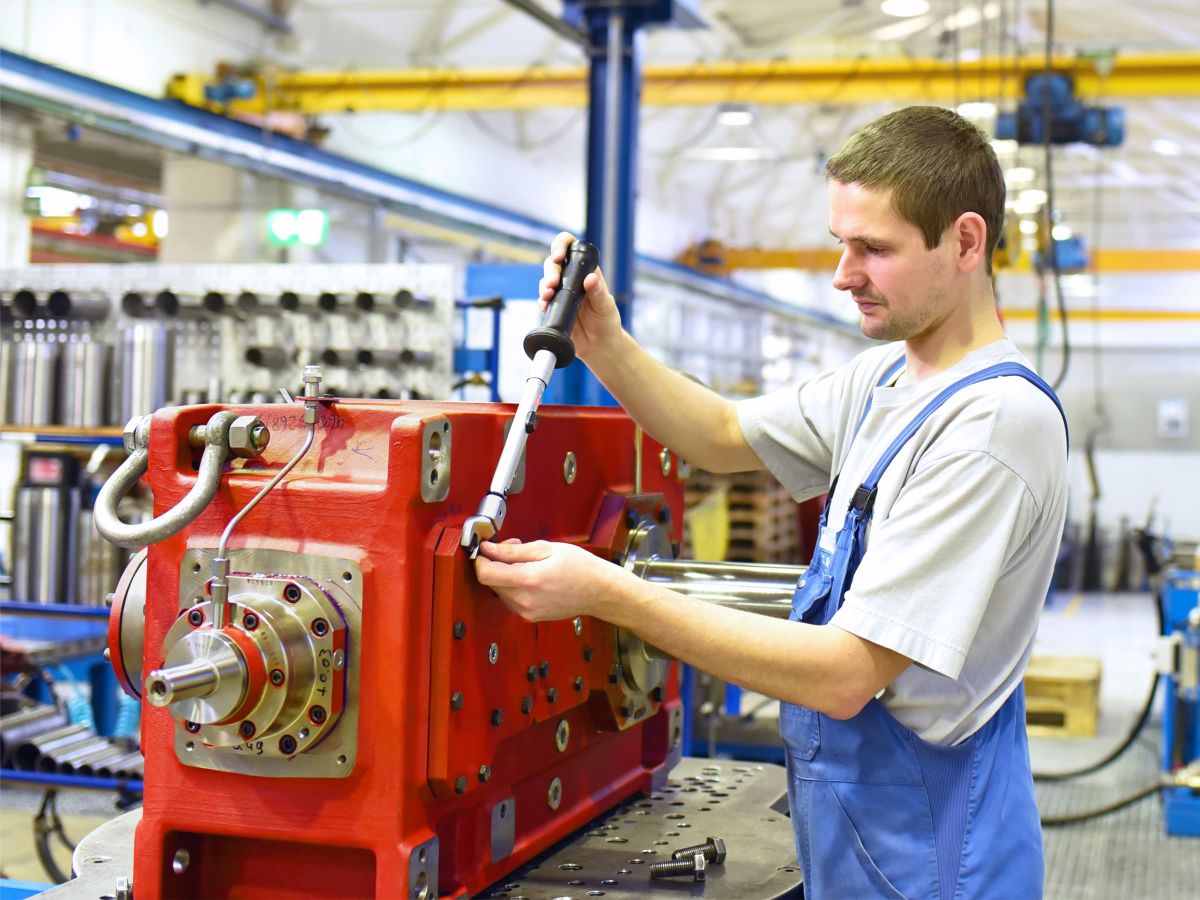 Ein Montagearbeiter in blauer Arbeitskleidung verwendet ein Werkzeug, um eine große rote Maschine in einer Produktionshalle zu reparieren.
