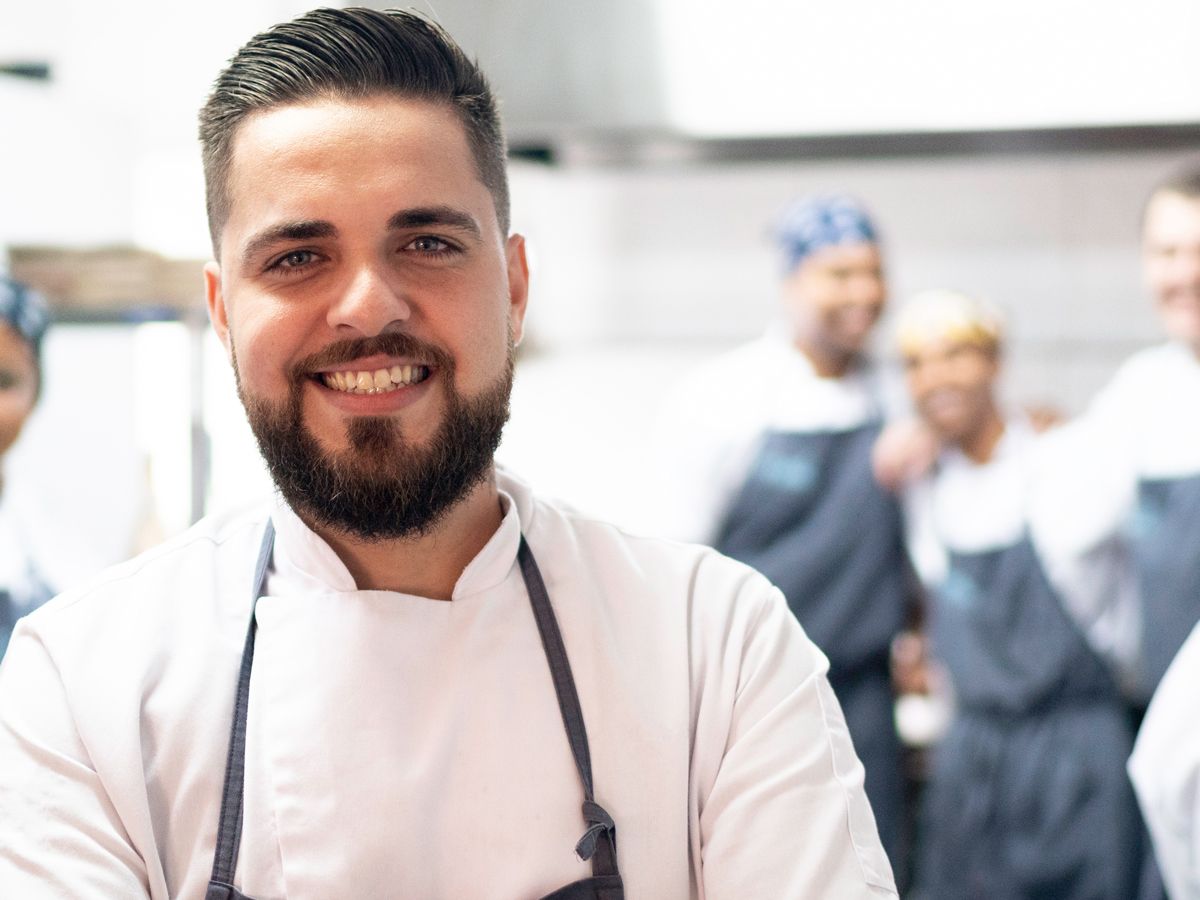 Ein lächelnder Koch in weißer Uniform steht im Vordergrund, während ein Team von Köchen unscharf im Hintergrund zu sehen ist.