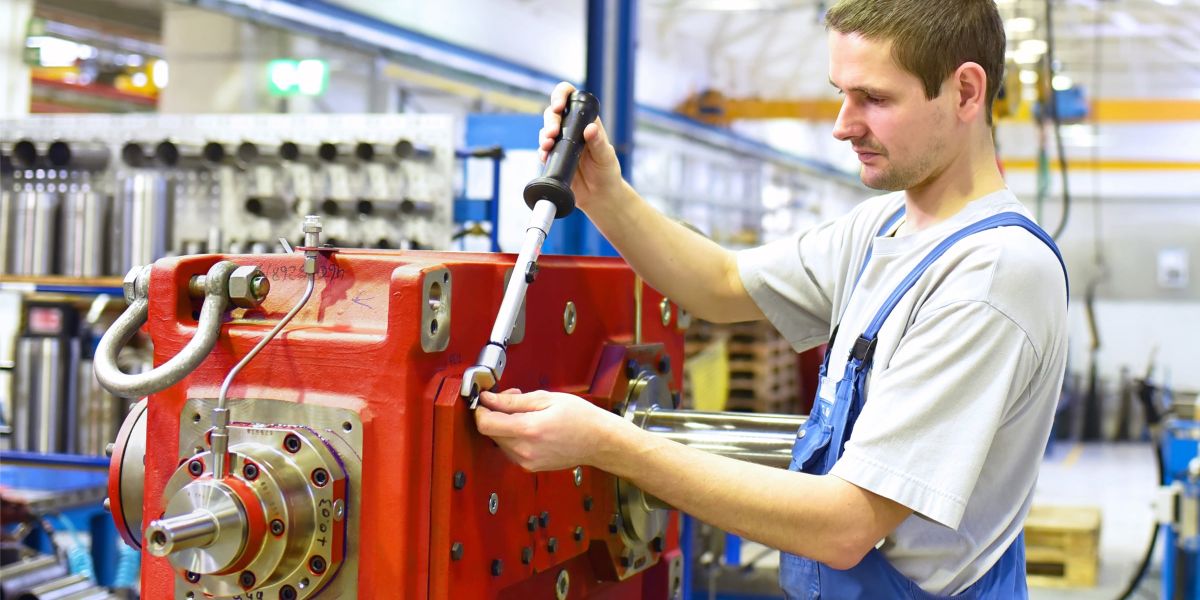 Ein Montagearbeiter in blauer Arbeitskleidung verwendet ein Werkzeug, um eine große rote Maschine in einer Produktionshalle zu reparieren.