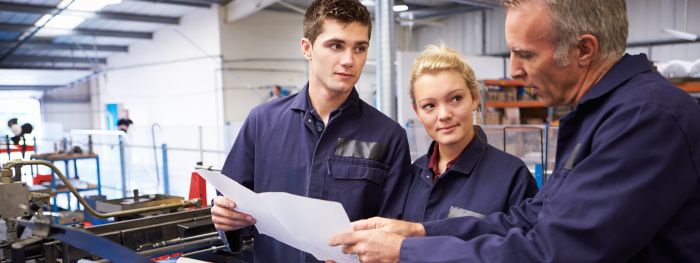 Ein Industriemeister erklärt zwei jungen Mitarbeitern in blauen Overalls einen Arbeitsprozess in einer Produktionshalle.