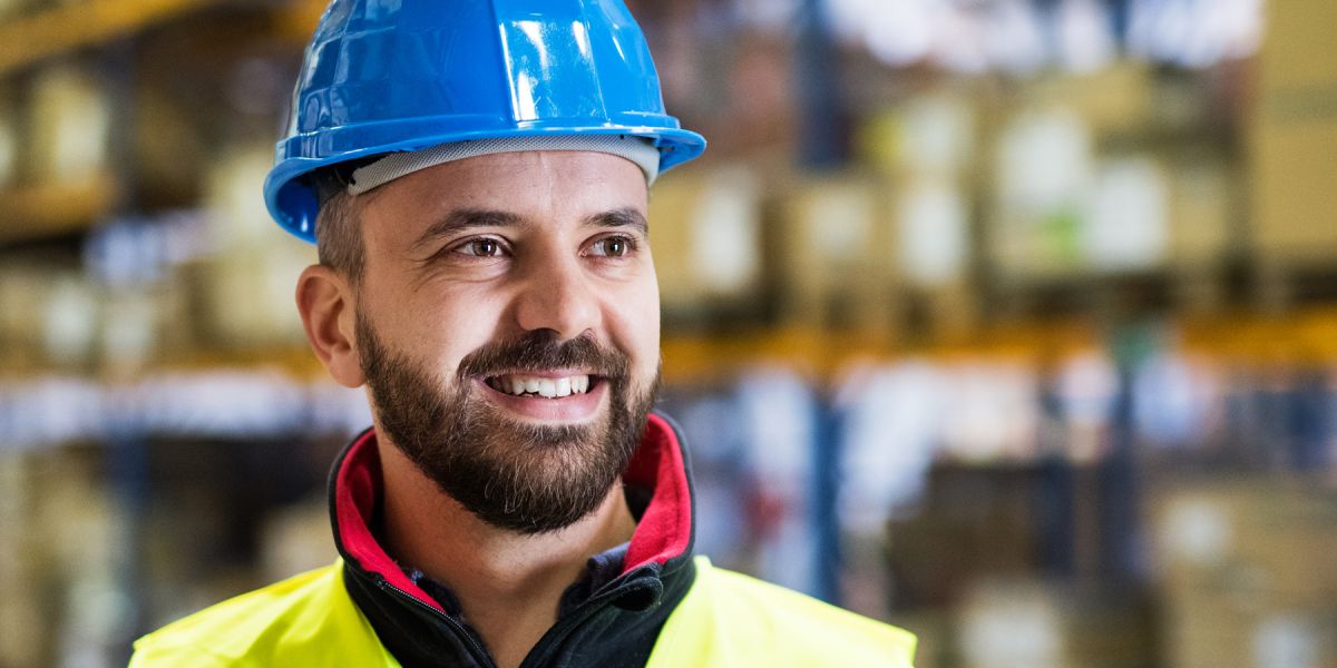 Lagermitarbeiter steht mit Schutzkleidung und Helm vor einem Hochregal und lächelt.